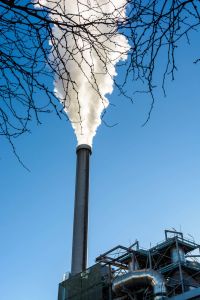 The tall metallic chimney of an incineration plant spitting out a heavy cloud of white smoke against a deep blue sky with tree branches in the foreground.